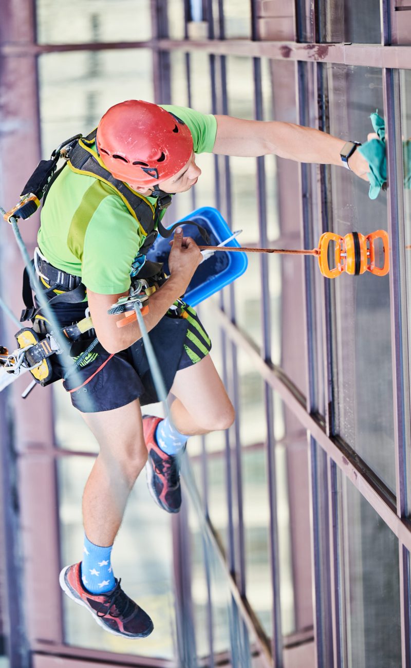 Industrial mountaineering worker washing glass windows of high-rise building, hanging on safety climbing rope. Man window cleaner in protective helmet cleaning skyscraper facade. Top view.